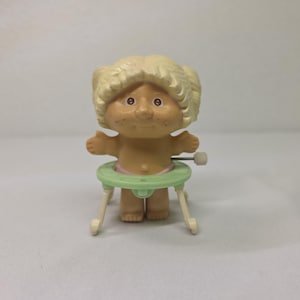 May include: A vintage doll with blonde hair and freckles, standing in a green and pink baby walker. The doll has its arms outstretched and a wind-up mechanism on the side. The toy is made of plastic.
