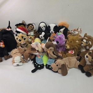 Op de afbeelding: Een grote verzameling diverse Ty Beanie Baby pluche speelgoed met verschillende dieren, waaronder een aap, een leeuw, een uil, een hert en een vlinder. Elk pluche dier heeft grote plastic ogen en een klein rood hartvormig Ty-label bevestigd.