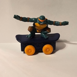 May include: A Teenage Mutant Ninja Turtle action figure in a skateboarding pose. The figure is green with an orange mask, brown accents, and a yellow shell. It is on a dark blue skateboard with orange wheels.