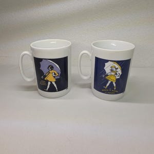 May include: Two white ceramic mugs featuring a graphic of a child in a yellow dress holding a white umbrella. The design includes the text "WHEN IT RAINS IT POURS" in white lettering against a dark blue background on each mug, with a handle on the side.