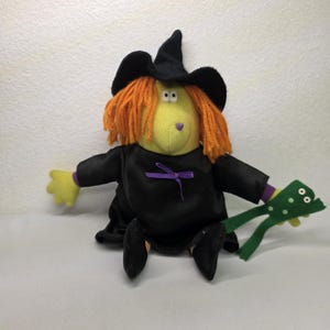 May include: A plush witch doll with a yellow face, orange yarn hair, and a black witch hat and dress. The doll has a purple bow and is holding a green frog. The doll's arms and hands are yellow.