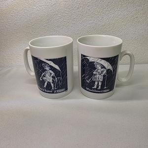 May include: Two white ceramic mugs featuring a dark blue graphic illustration of a child holding an umbrella in the rain. Below the illustration, the text reads When It Rains IT POURS. Each mug has a curved handle and a smooth, glossy finish.