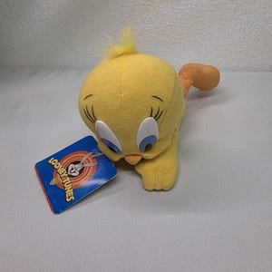 May include: A yellow plush Tweety Bird toy with blue eyes, orange feet, and a tuft of yellow hair. The Looney Tunes tag is attached. The plush toy is lying on its stomach.