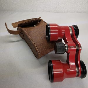 May include: Red opera glasses with black accents and a brown leather case. The vintage binoculars have a central focus knob and are in a closed position. The case has a flap closure and a small handle.