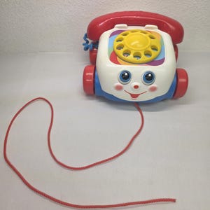 May include: A vintage pull-along toy phone in red, white, blue, and yellow. The toy has a smiling face, rolling wheels, and a rainbow dial. A red pull cord extends from the front.