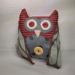 May include: A handmade owl-shaped pillow with a red and gray color scheme. The owl has large, round eyes with red button centers, a cream-colored beak, and wings with a leaf pattern. A large yellow button is on the owl's chest.