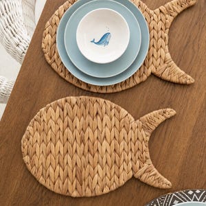 Peut inclure: Deux sets de table tressés en forme de poisson, de couleur marron clair et en fibres naturelles. L'un des sets supporte une pile de deux assiettes bleu clair et un bol blanc orné d'une petite illustration de baleine bleue.