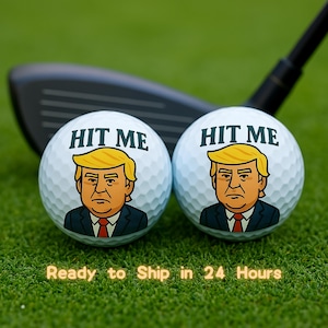May include: Two white golf balls with a cartoon image of a person with blonde hair and a serious expression. The text "HIT ME" is printed on each ball.