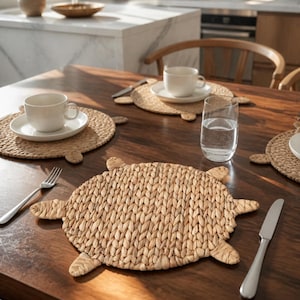 Peut inclure: Un ensemble de sets de table ronds en jonc de mer tressé en forme de tortue, avec une texture nattée et de petits membres saillants. Ces sets de table de couleur naturelle sont disposés sur une table à manger en bois foncé avec des tasses.