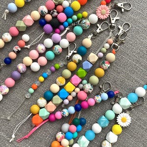 Puede incluir: Una colección de clips para chupete con cuentas de colores de varias formas y tamaños. Los clips presentan una variedad de colores, incluyendo rosa, azul, amarillo y verde, con cierres de metal. Algunas cuentas tienen diseños florales.