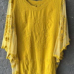 Könnte beinhalten: Senfgelbe Bluse mit aufwendiger floraler Stickerei und transparenten Glockenärmeln. Das Oberteil hat einen wellenförmigen Saum und einen detaillierten Ausschnitt. Der Stoff wirkt leicht. Die Bluse hängt an einem weißen Kleiderbügel.