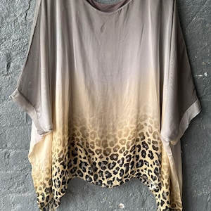 May include: A draped top with a gradient design, transitioning from light grey to beige, and featuring a leopard print hem. The top has short, wide sleeves and is hanging on a black hanger against a grey brick wall.