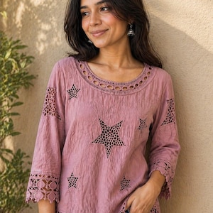 Puede incluir: Blusa rosa empolvado con recortes de estrellas y ribete de encaje. La parte superior de manga larga presenta un escote redondo con un diseño calado decorativo. La blusa se combina con unos vaqueros azules.