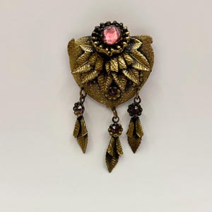 May include: Vintage-style brooch with a heart-shaped base and intricate leaf detailing. The brooch features a central pink gemstone surrounded by smaller gems and dangling leaf-shaped pendants. The metal has a warm, antique gold finish.