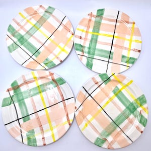 May include: A set of four round ceramic plates featuring a hand-painted plaid pattern. The design consists of intersecting brushstrokes in shades of peach, sage green, bright yellow, and thin black lines against a white background surface.