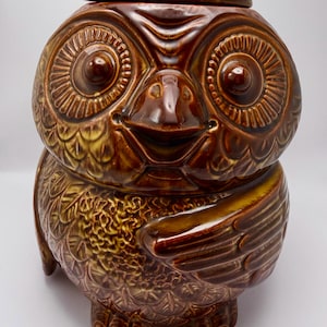 May include: A ceramic owl-shaped cookie jar with a lid. The jar is primarily brown with yellow and tan accents. The owl has large, detailed eyes, a smiling beak, and wing details. The jar is a decorative kitchen item.