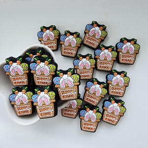 May include: A collection of 16 colourful Easter-themed charms featuring bunnies in baskets with the text "Happy Easter".