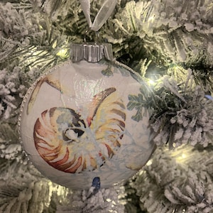 May include: A white Christmas ornament with a seashell design. The ornament has a white background with a gold and red seashell in the center. The seashell is surrounded by a blue and white design.