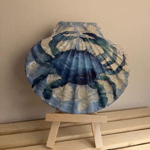 May include: A blue crab painted on a white seashell with a blue and green watercolor wash. The seashell is displayed on a small wooden easel.