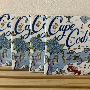 May include: Set of four white ceramic coasters with a blue and white illustration of Cape Cod, featuring a lighthouse, beach chairs, a lobster, and the text "Cape Cod".