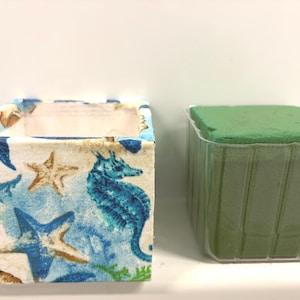 May include: A square container featuring a nautical pattern of blue seahorses, tan starfish, and coral on a white background, positioned next to a clear plastic insert filled with a solid green block of floral foam for arrangement support.
