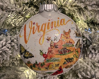May include: A white Christmas ornament with a map of Virginia, featuring a butterfly, cardinal, and the word "Virginia" in orange script. The ornament is hanging on a frosted Christmas tree.