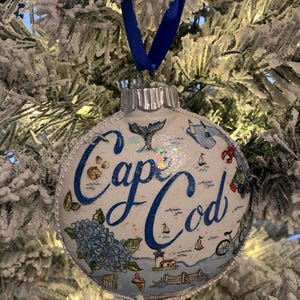 May include: White Christmas ornament with a blue and white illustration of Cape Cod, featuring a lighthouse, a bicycle, and a whale tail. The ornament has a silver trim and a blue ribbon.