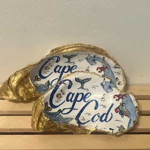 May include: Two oyster shells with gold-painted edges. The shells are decorated with a map design and the words "Cape Cod" in blue script. The map includes illustrations of whales, sailboats, and lobsters.