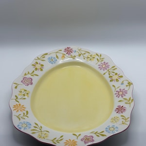 Op de afbeelding: Een decoratieve, geschulpte bord met een lichtgele kern en een witte rand. De rand is versierd met een bloemenpatroon in tinten roze, blauw, geel en oranje. Het bord heeft een donkerrode rand.