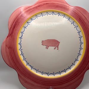 May include: A decorative ceramic plate with a scalloped edge. The plate features a red rim, a yellow band, and a white center with a hand-painted pig illustration. Blue accents add detail to the design. The plate is a charming addition to any kitchen.