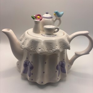 以下が含まれることがあります： 白い陶器製のティーポット。蓋にはフリルがあしらわれ、ミニチュアのティーポット、ティーカップ、ピンクの花が活けられた小さな花瓶が飾られています。ポット本体には紫色の水玉模様と、底の周りに紫色のリボンが描かれています。