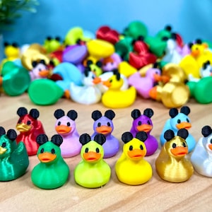 May include: A collection of small, multi-colored plastic duck figurines, each featuring a pair of black mouse-like ears on top of their heads. The ducks are rendered in various solid colors including green, red, pink, purple, yellow, gold, and white, all with orange beaks.