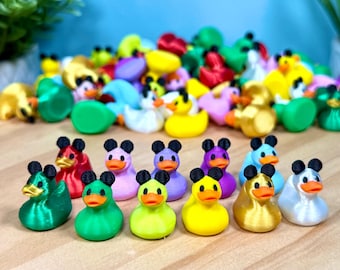 Puede incluir: Una colección de pequeñas figuras de patos de plástico multicolores, cada una con un par de orejas negras similares a las de un ratón en la parte superior de la cabeza. Los patos son de varios colores sólidos, incluyendo verde, rojo, rosa, morado, amarillo, dorado y blanco, todos con picos naranjas.