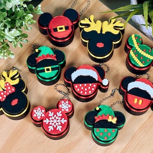 May include: A collection of nine circular keychains featuring mouse-ear silhouettes with various holiday designs, including Santa suits, reindeer antlers, elf hats, Christmas trees, and snowflakes in red, green, black, gold, and white plastic.