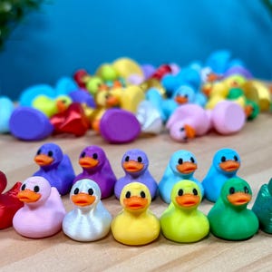 May include: A collection of small, multi-colored rubber ducks arranged in rows. The ducks feature vibrant shades including red, pink, white, yellow, green, blue, and purple, each with a distinct orange beak and black eyes.