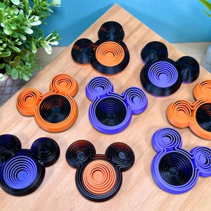 Op de afbeelding: Een verzameling van zeven 3D-geprinte fidget-speeltjes in de vorm van muizenkoppen, met concentrische cirkelpatronen. De speeltjes zijn gekleurd in verschillende combinaties van zwart, oranje en paars, gerangschikt op een licht houten oppervlak.