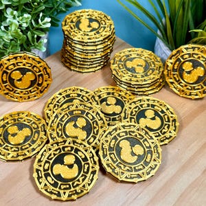 Könnte beinhalten: Eine Sammlung runder goldener und schwarzer Spielmarken mit einer erhabenen Mickey-Mouse-Silhouette in der Mitte. Die Spielmarken haben ein kompliziertes Randdesign mit geometrischen Mustern und sind auf einer hellen Holzoberfläche angeordnet.