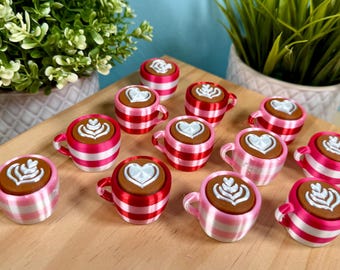 Könnte beinhalten: Eine Sammlung von Miniatur-Kaffeetassen-Dekorationen. Jede Tasse hat eine braune Oberseite mit weißer Latte-Art und einen rosa-weiß gestreiften Körper. Die Tassen sind auf einer Holzoberfläche angeordnet.