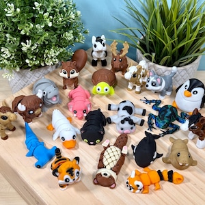 Pode incluir: Uma coleção de figuras de animais coloridas, incluindo um esquilo, um pinguim, um tigre e um sapo, dispostas sobre uma superfície de madeira. As figuras são feitas de um material macio e são de vários tamanhos. Plantas verdes no fundo.