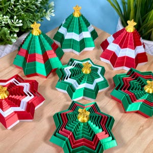 以下が含まれることがあります： 赤、緑、白の折りたたみ式クリスマスツリーオーナメント7個セット。それぞれ小さな金色の星が頂点についています。これらの幾何学的な段状の装飾品は木製の表面に配置されており、直立した状態と平らな状態の両方が見られます。