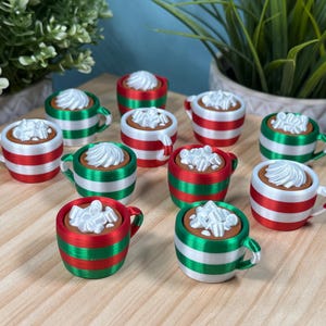 May include: A collection of miniature hot chocolate mugs with whipped cream topping. The mugs are decorated with red, white, and green stripes, and are arranged on a light wood surface. The scene evokes a festive holiday theme.