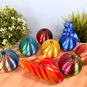 May include: A collection of colorful, spherical, and abstract 3D-printed objects. The objects are in various colors, including blue, green, red, and gold. Some are assembled, while others are disassembled, revealing their internal structure. The objects are arranged on a wooden surface.