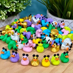 May include: A large collection of miniature rubber ducks in various colors including yellow, pink, green, blue, purple, and metallic gold. Each small duck features a distinct orange beak and two black circular ears on top of its head.