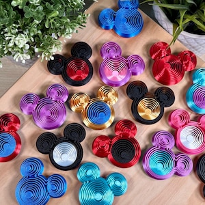 Puede incluir: Una colección de decoraciones en forma de Mickey Mouse con diseño en espiral y colores vibrantes. Las decoraciones son rojas, azules, negras, doradas y moradas, dispuestas sobre una superficie de madera marrón claro, con vegetación de fondo.