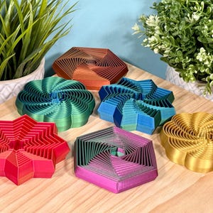 May include: A collection of colorful, geometric 3D-printed objects in various shapes, including stars, hexagons, and spirals. The objects feature gradient color schemes of red, green, blue, and gold. They are displayed on a light wood surface.