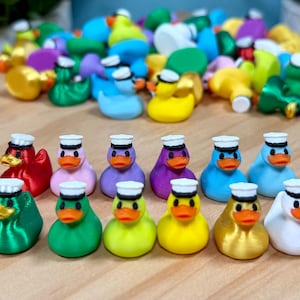 May include: A collection of miniature rubber ducks in various solid colors, each wearing a small white sailor hat with a black band. The ducks feature orange beaks and black eyes, arranged in a neat row in the foreground with a pile of additional ducks behind.