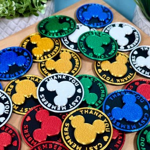 May include: A collection of circular plastic pins featuring a raised Mickey Mouse head silhouette in the center. Each pin has a black background with the text THANK YOU CAST MEMBERS printed in white, blue, green, red, or gold metallic colors.