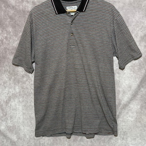 Puede incluir: Un polo de manga corta con un patrón texturizado en tonos grises, marrones y azules. El cuello es negro con una fina raya roja y blanca. La camisa tiene una tapeta de tres botones y la marca "Penguin" es visible en el cuello.