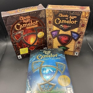 May include: Three PC game expansion packs for "Dark Age of Camelot". The boxes feature the game title and artwork, with titles including "Catacombs", "Shrouded Isles", and "Trials of Atlantis". The boxes are in shades of brown, gold, and blue.