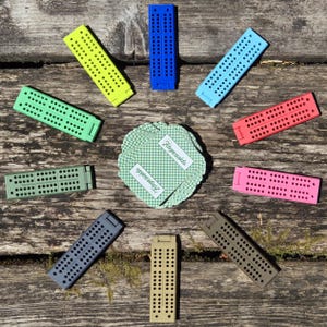 May include: A collection of colorful cribbage boards in various colors, including yellow, blue, green, and pink, arranged around a deck of cards with the text "Fingeraids". The boards are made of plastic and have numerous holes for pegs.
